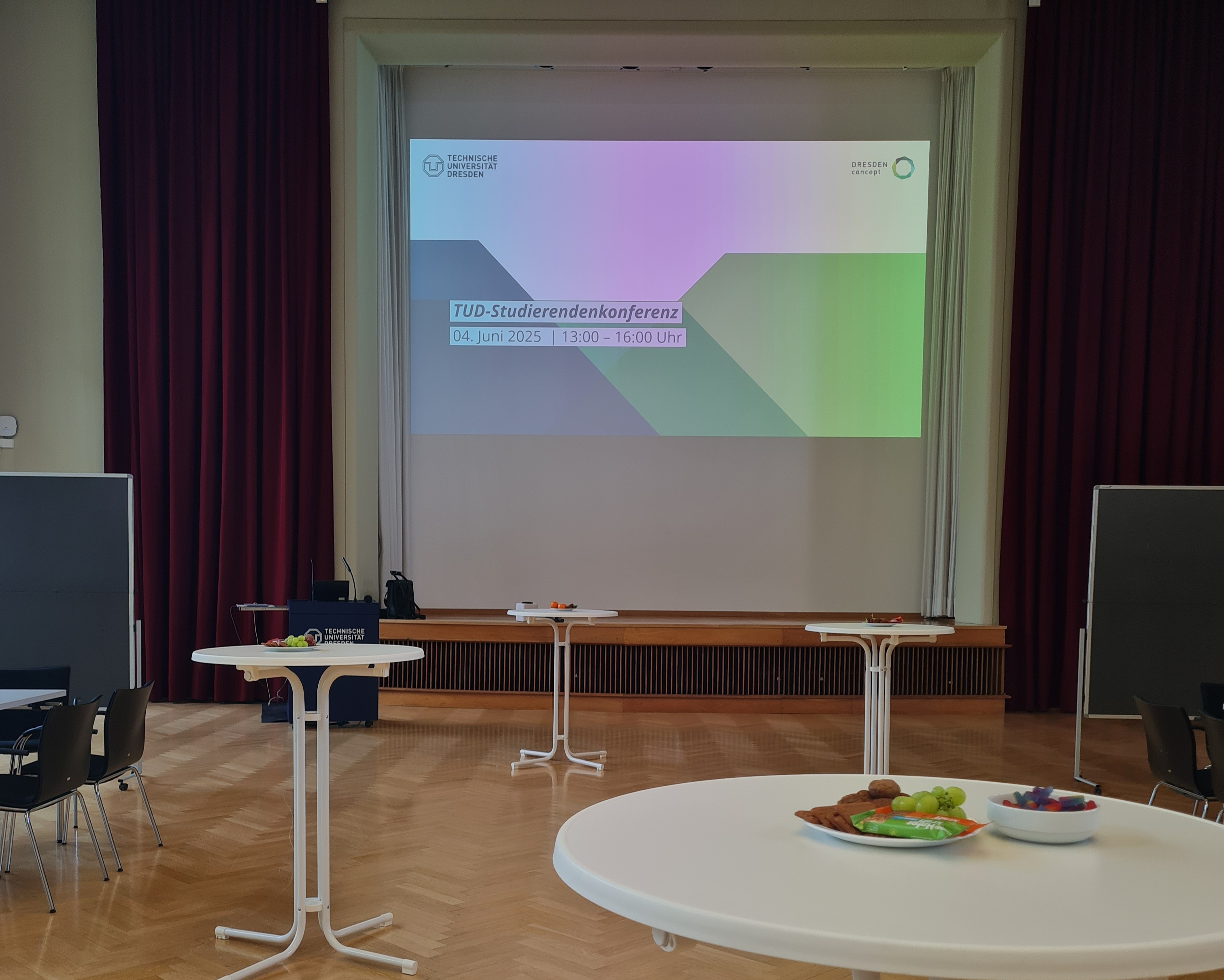 Der Dülfersaal mit mehreren Stehtischen, im Hintergrund ist eine Präsentation angebeamt mit dem Titel TUD Studierendenkonferenz