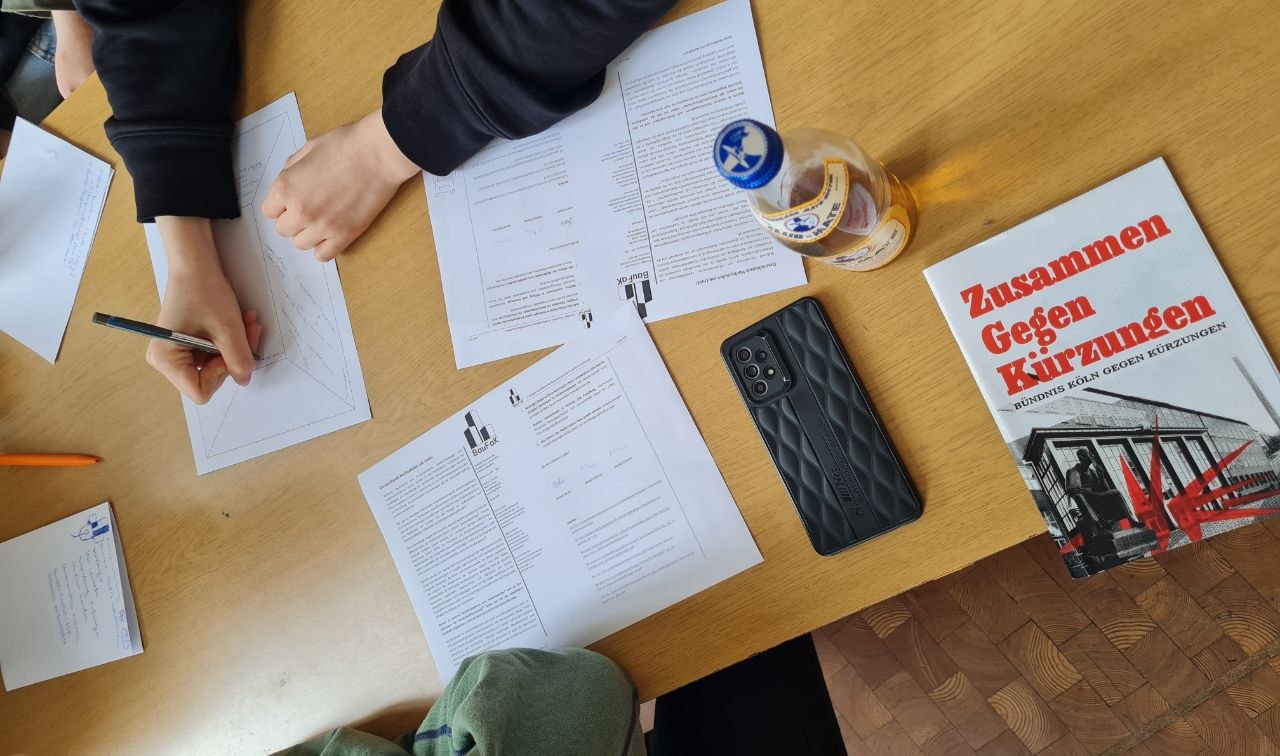 Gruppenarbeit Kürzungen Das Bild zeigt einen Tisch von oben, an dem mehrere Leute sitzen und gemeinsam arbeiten. Es wird etwas aufgeschrieben und es liegen Texte und eine Broschüre zu Kürzungen auf dem Tisch.