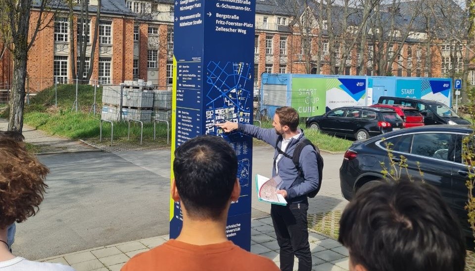 Eine Gruppe Studierender steht vor einer Säule des Campusleitsystem. Der Campusführer erklärt etwas und zeigt auf die Säule.