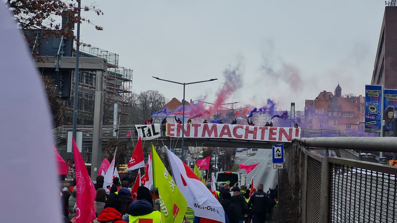 Die Fußgängerbrücke über der Bergstraße zum HSZ, darunter läuft eine Demo, auf der Brücke ist ein großes Banner gespannt mit der Schrift "TdL entmachten"