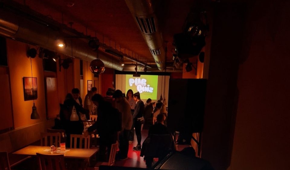 Im Club Aquarium ist eine Menge an Studierender zu sehen. Im Hintergrund wird ein Pub Quiz an die Wand projiziert.