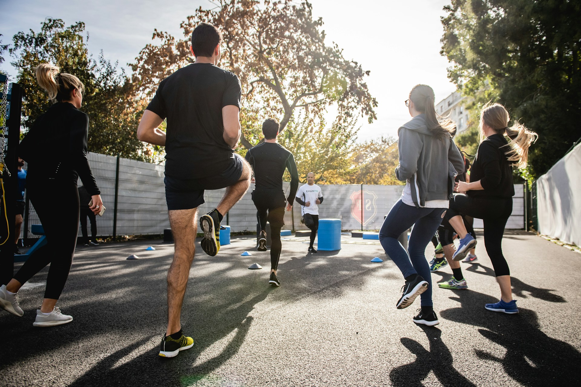 Group Fitness Training im Freien Mehrere Personen beim Aufwärmtraining im Freien von hinten gesehen. Sie absolvieren Lauf- und Hüpfübungen auf einem geteerten Platz bei sonnigem Wetter.