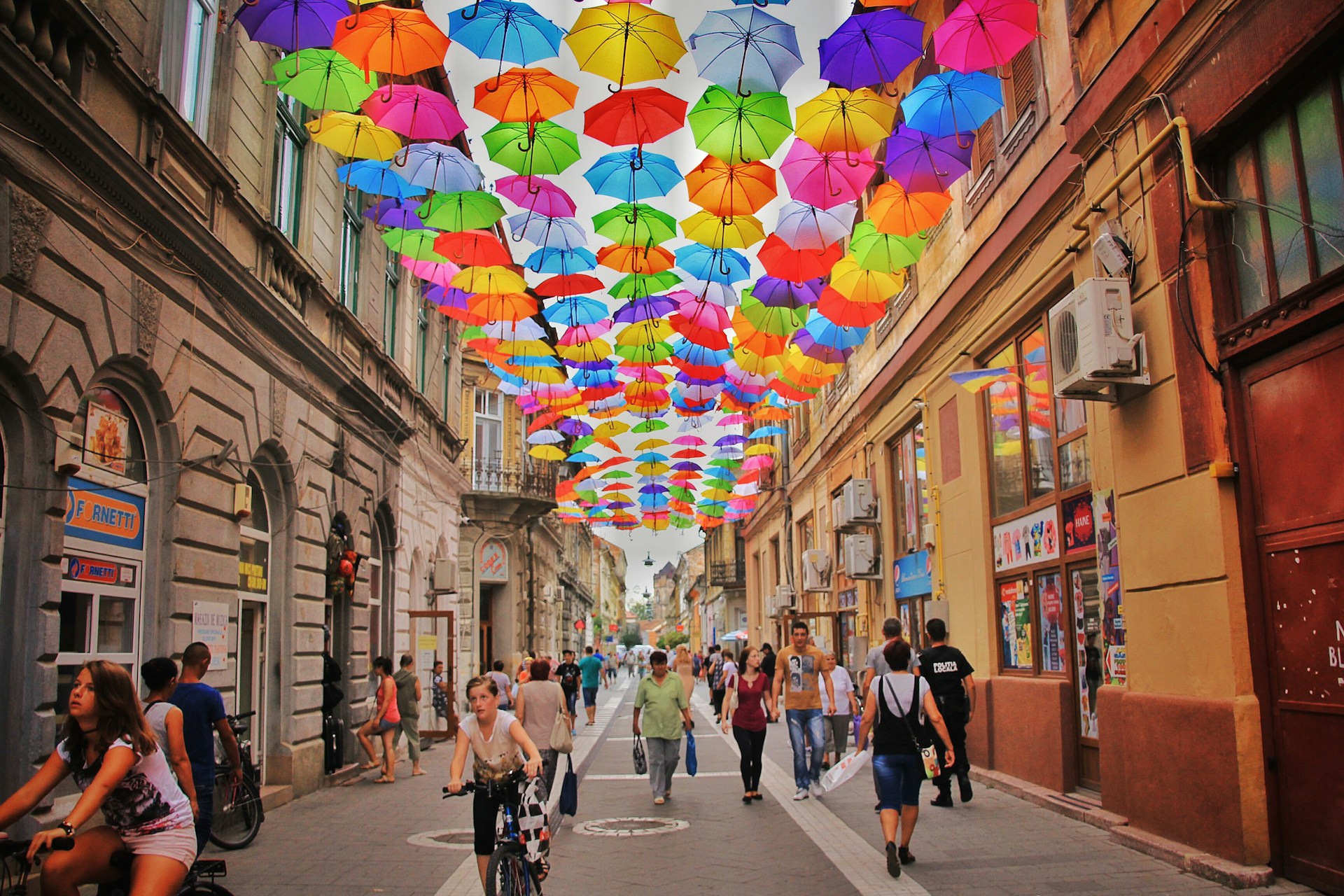 Bunter Regenschirm-Himmel in der Altstadt Zahlreiche bunte Regenschirme in Rot, Gelb, Blau und Grün hängen schwebend zwischen alten Gebäuden über einer Fußgängerzone mit vielen Passanten und Radfahrern.