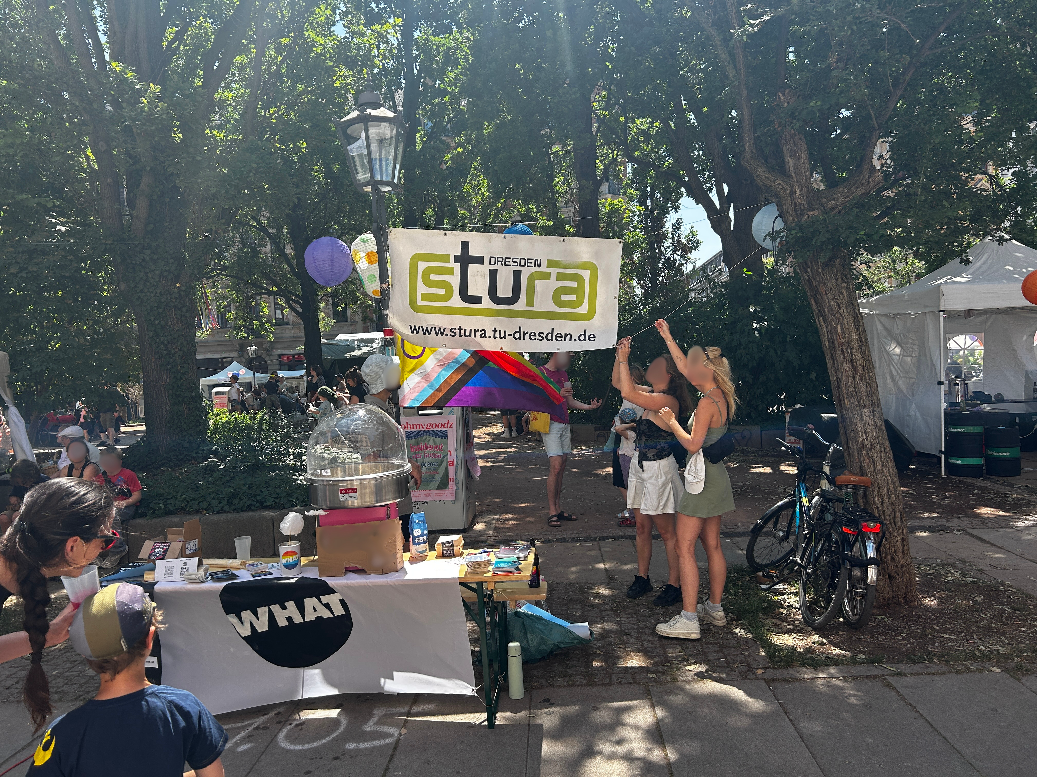 Sommerlicher Tag, Martin-Luther-Platz. Stand mit Zuckerwattemaschine im Vordergrund, Stura Banner hängt in den Bäumen im Hintergrund, WHAT Flagge hängt über Tisch, Menschen im Vordergrund