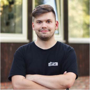 Bild zeigt Mensch mit StuRa Logo auf dem T-Shirt und verschränkten Armen lächelnd. Im Portrait Modus fotografiert vor der StuRa-Baracke